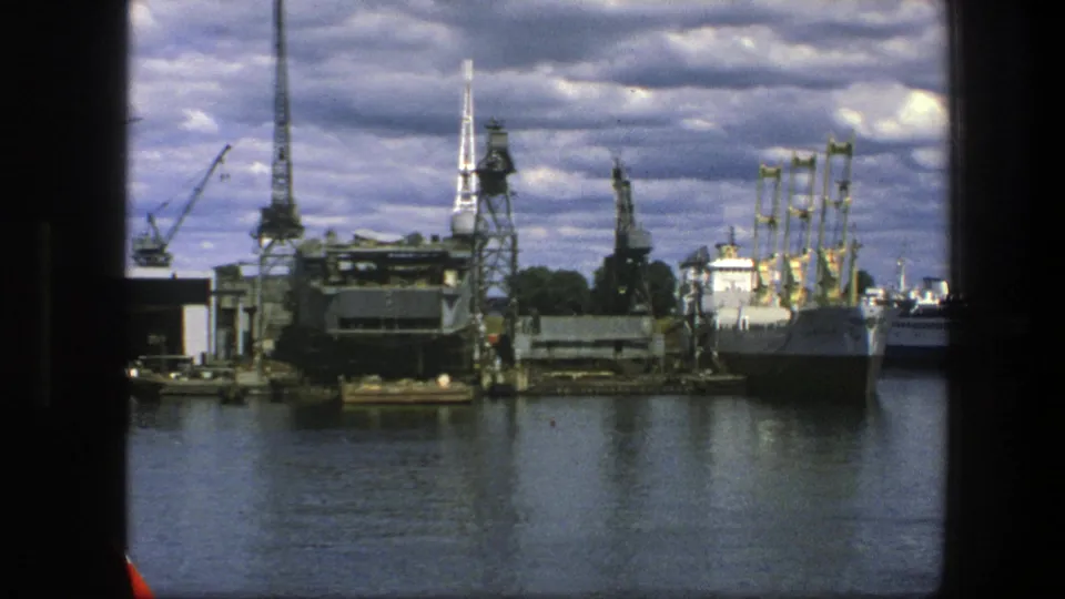 1975 Copenhagen harbor view with cranes, industrial shoreline buildings, dark water and a ship seen through a window edge.