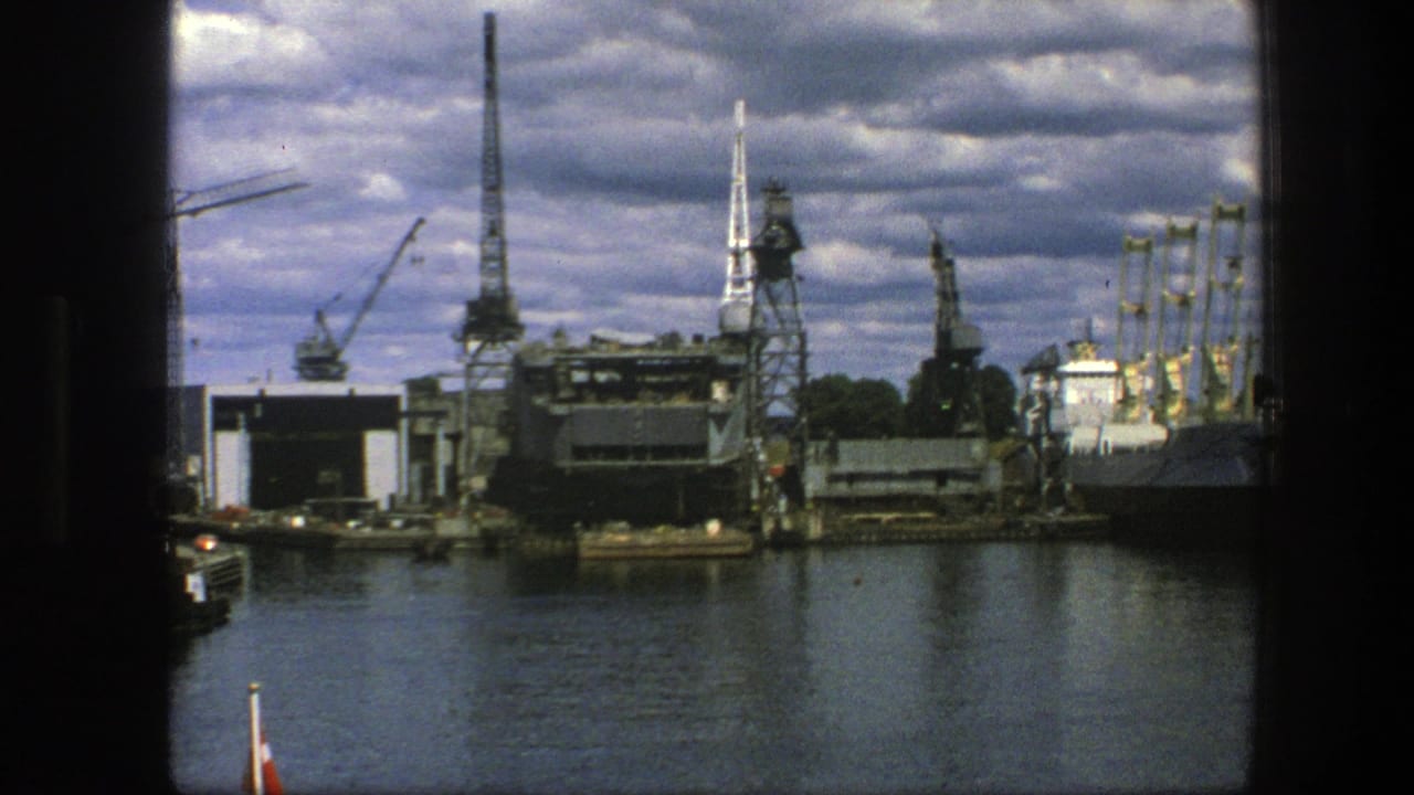 Copenhagen industrial waterfront in 1975, with cranes, factory-like structures and moored vessels under heavy clouds.