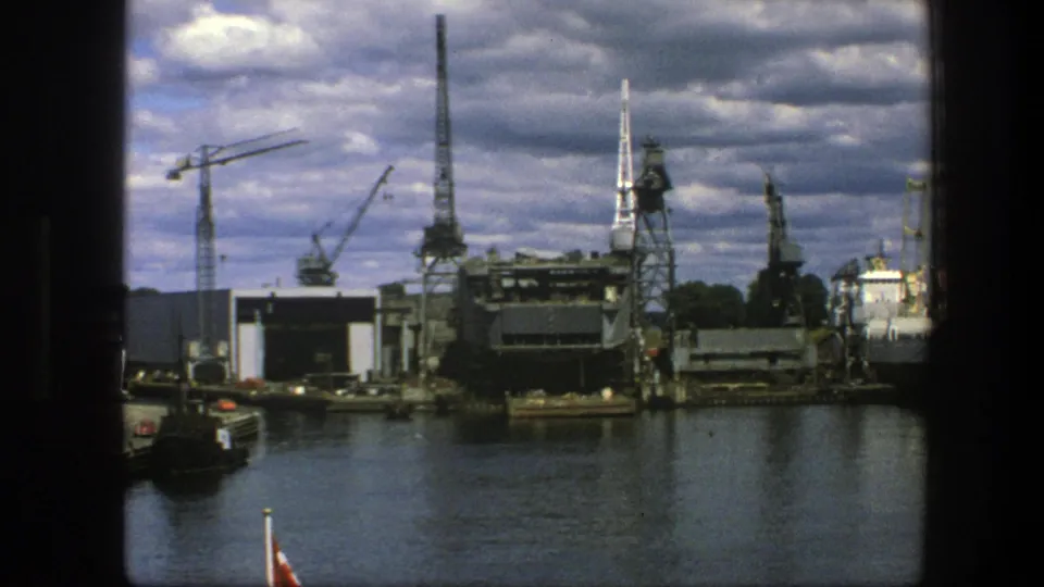 1975 Copenhagen port scene with cranes, industrial sheds, dark water and a small boat entering the lower-left frame.