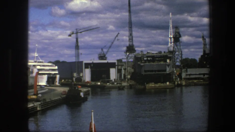 1975 Copenhagen harbor view with a passenger ship at a pier, industrial cranes, dark water, and a cloudy sky seen through a frame edge.