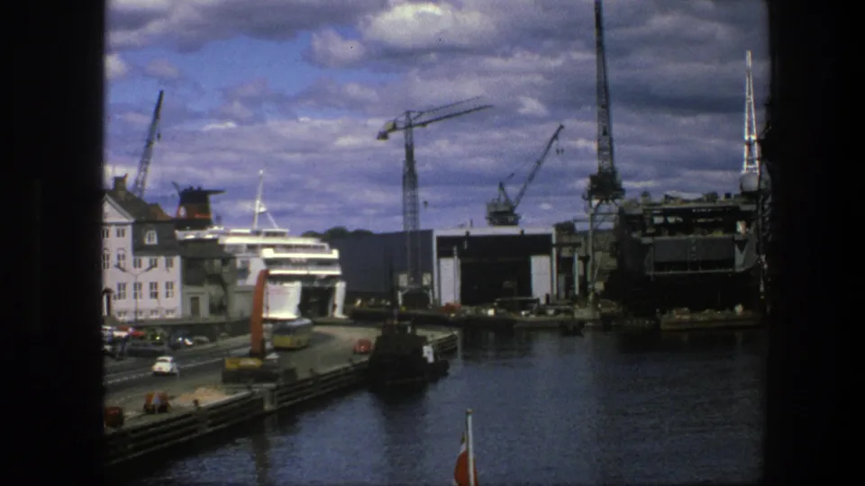 1975 Copenhagen waterfront view with a road and cars, a pale harbor building, a docked passenger ship, cranes, and dark harbor water.