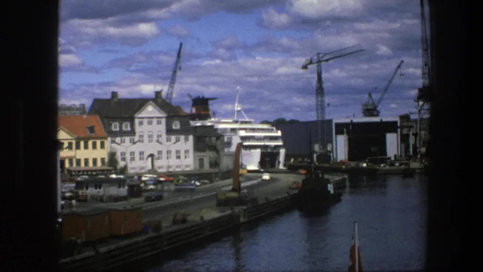 1975 Copenhagen harbor frame showing waterfront buildings, a large white ship, cranes, cars on the quay, and dark edges from the filming position.