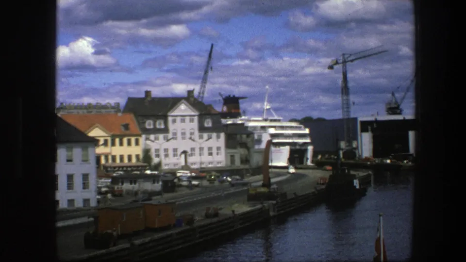 Late frame of the 1975 Copenhagen harbor view with a white passenger ship, cranes, waterfront road traffic, buildings, and reflective water.