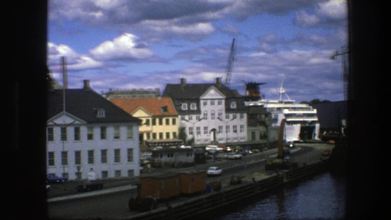 Harbor road, white buildings, docked ship, cranes, water, and scattered clouds.