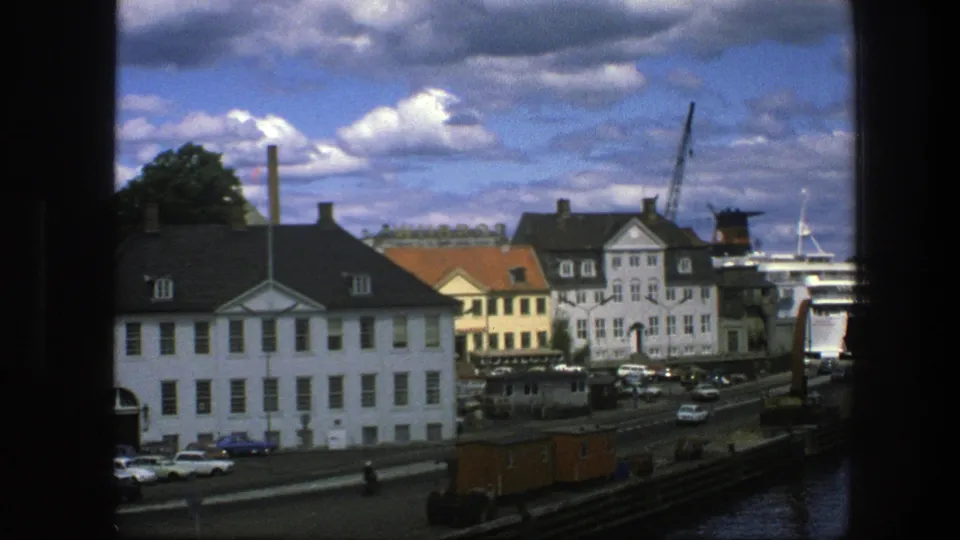 1975 Copenhagen waterfront view with a prominent white building in front, cars on the road, cranes, a partially visible ship, harbor water, and clouds.