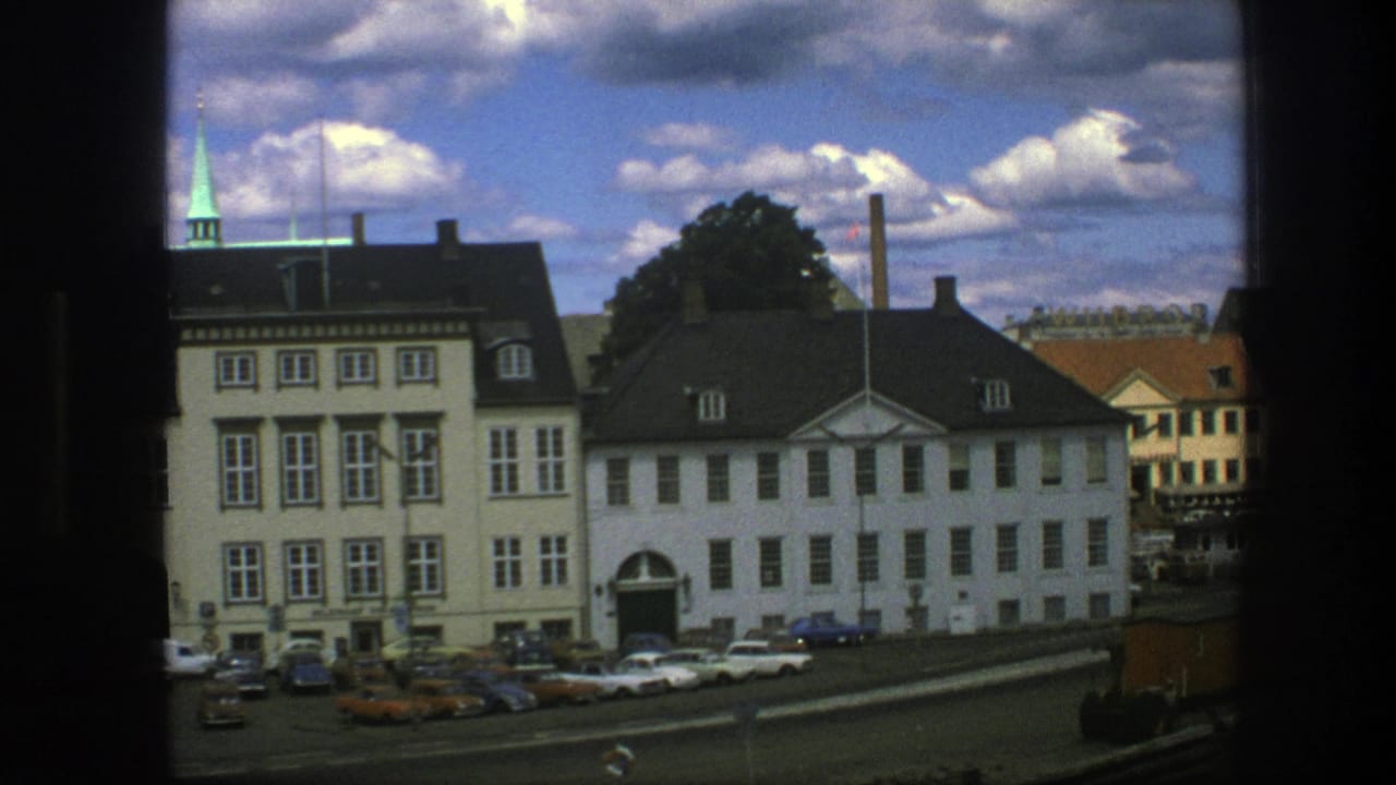 1975 Copenhagen view of pale facades, parked cars, a distant spire, and a partly cloudy blue sky from a window-framed angle.