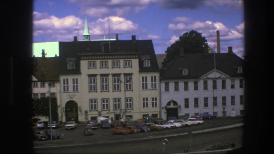 1975 Copenhagen street view with multi-story pale buildings, dark roofs, parked cars, a spire, and a visible construction crane.