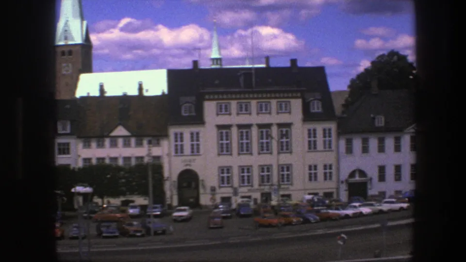 1975 Copenhagen 8mm frame showing pale buildings, street traffic, parked cars, a tall church tower, and dark window-frame borders.