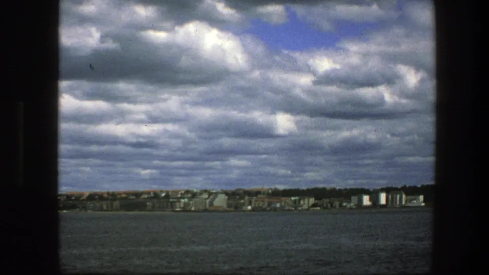Copenhagen shoreline in the distance across dark water, filmed from a boat under layered gray clouds with small blue breaks.