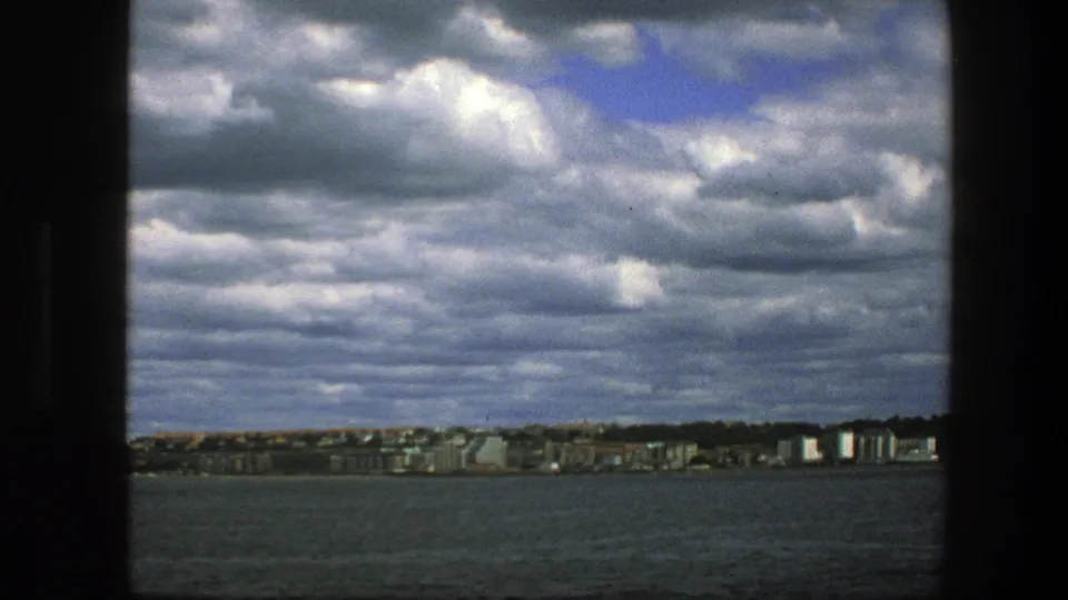 A 1975 boat passenger view across Copenhagen waters, showing a low urban waterfront beneath a dense, textured cloud ceiling.