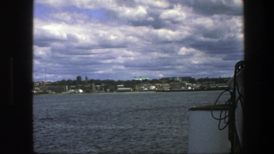 Copenhagen waterfront filmed from a boat, with choppy water, distant buildings, cloud cover, and part of the vessel visible at the right edge.