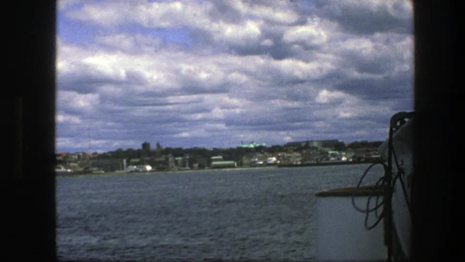 1975 home-movie frame of Copenhagen across the bay, with dramatic clouds and boat hardware at the edge.