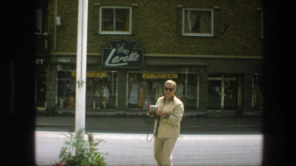 Street scene in Copenhagen with an adult man holding a camera near shop windows and a readable storefront sign.