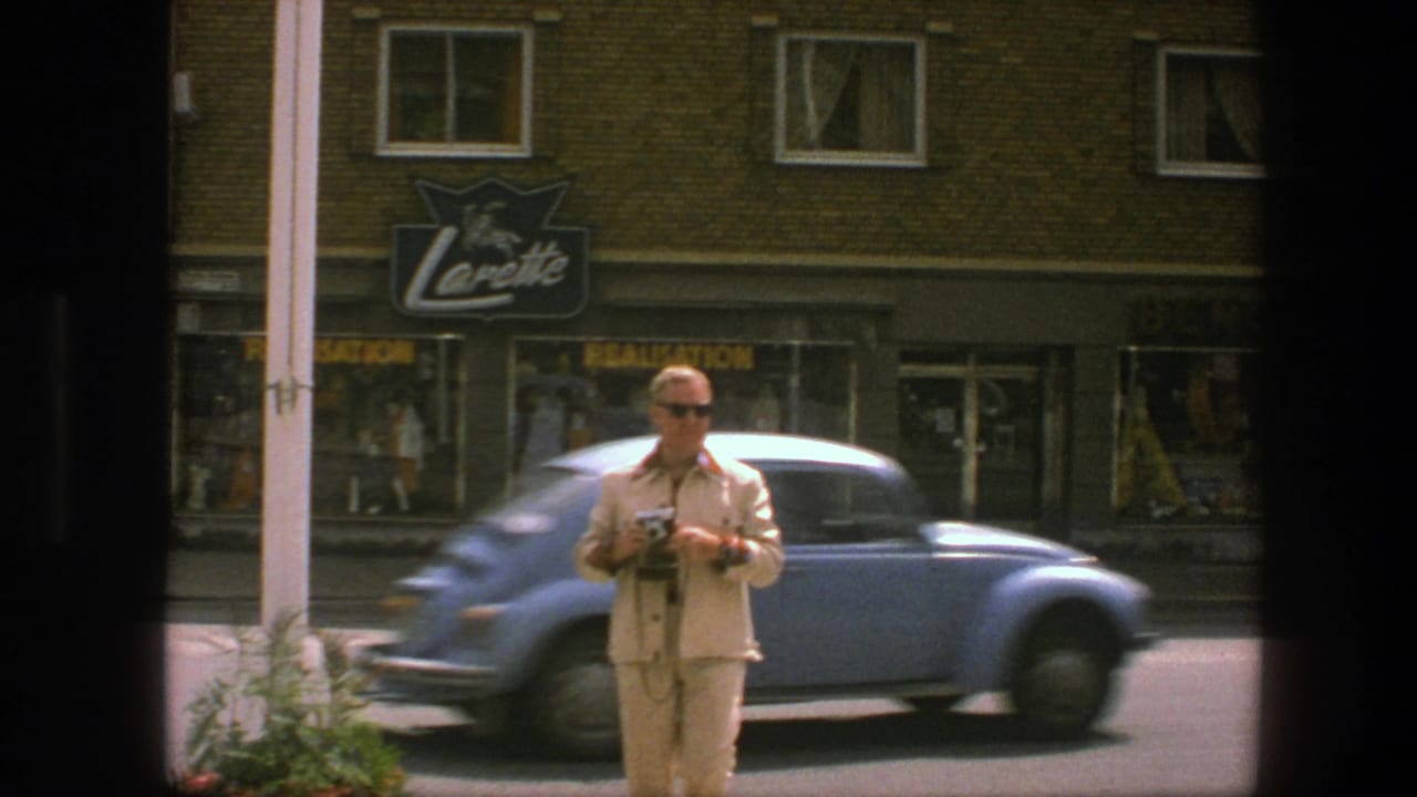 Man in a light suit holds a camera as a blue Volkswagen Beetle passes on a city street.