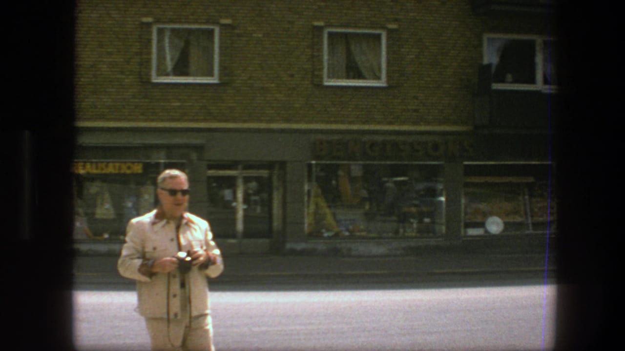 Man with sunglasses and camera stands in the street foreground near storefronts.