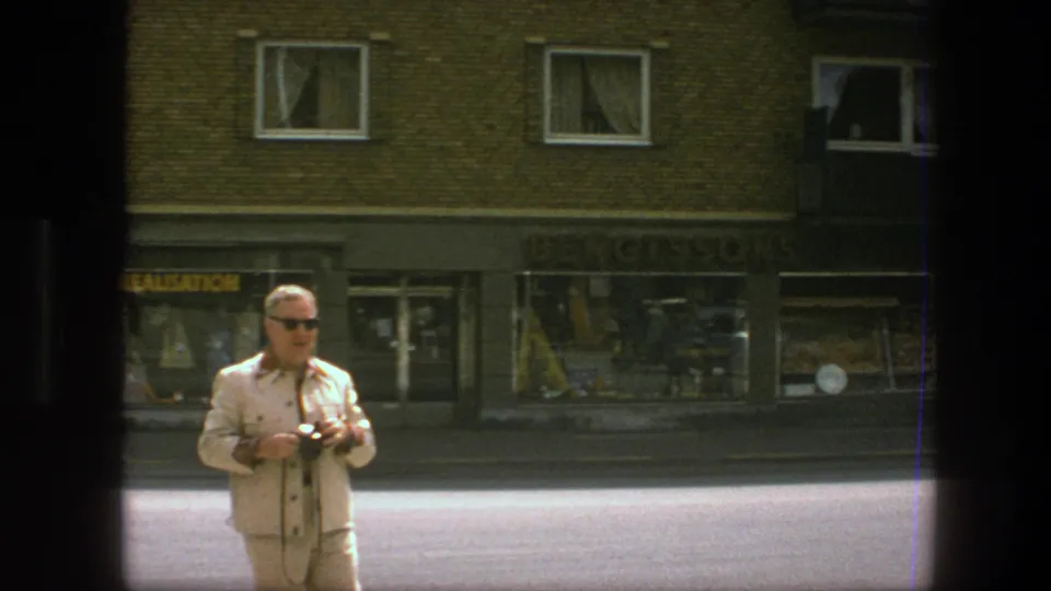 The man remains centered on a Copenhagen street, holding his camera with shop windows and apartment windows behind him.