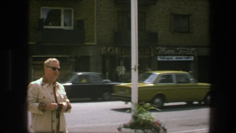 A man in a light suit holds a camera in the foreground while dark and yellow cars pass along a Copenhagen street in 1975.