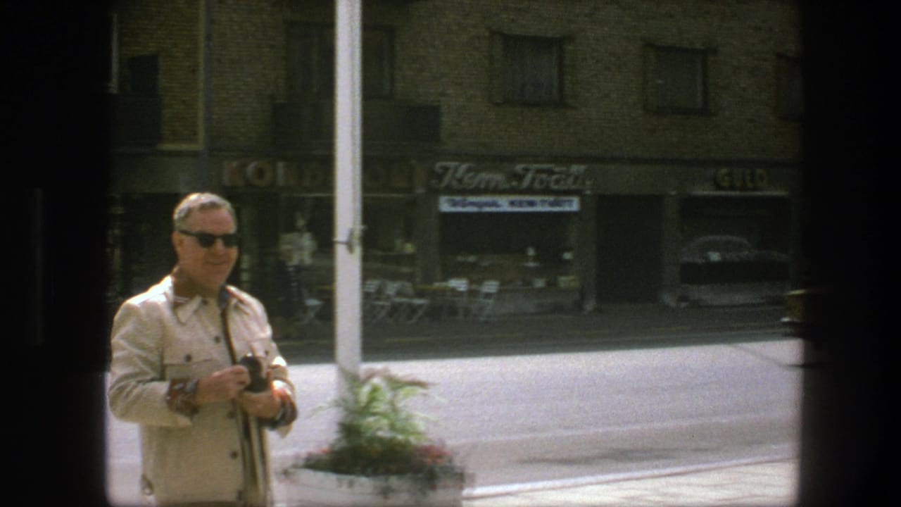 1975 Copenhagen street frame with a man holding a camera, brick apartment facade, shop signs, and outdoor seating across the road.