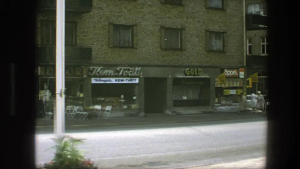 1975 Copenhagen street frame showing a row of small shops, apartment windows, outdoor chairs, and dark 8mm frame edges.