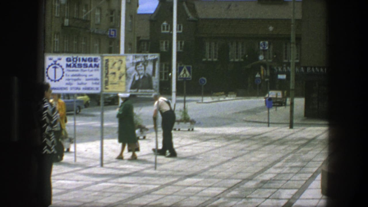 Pedestrians cross a paved plaza near advertising boards and storefront signage.