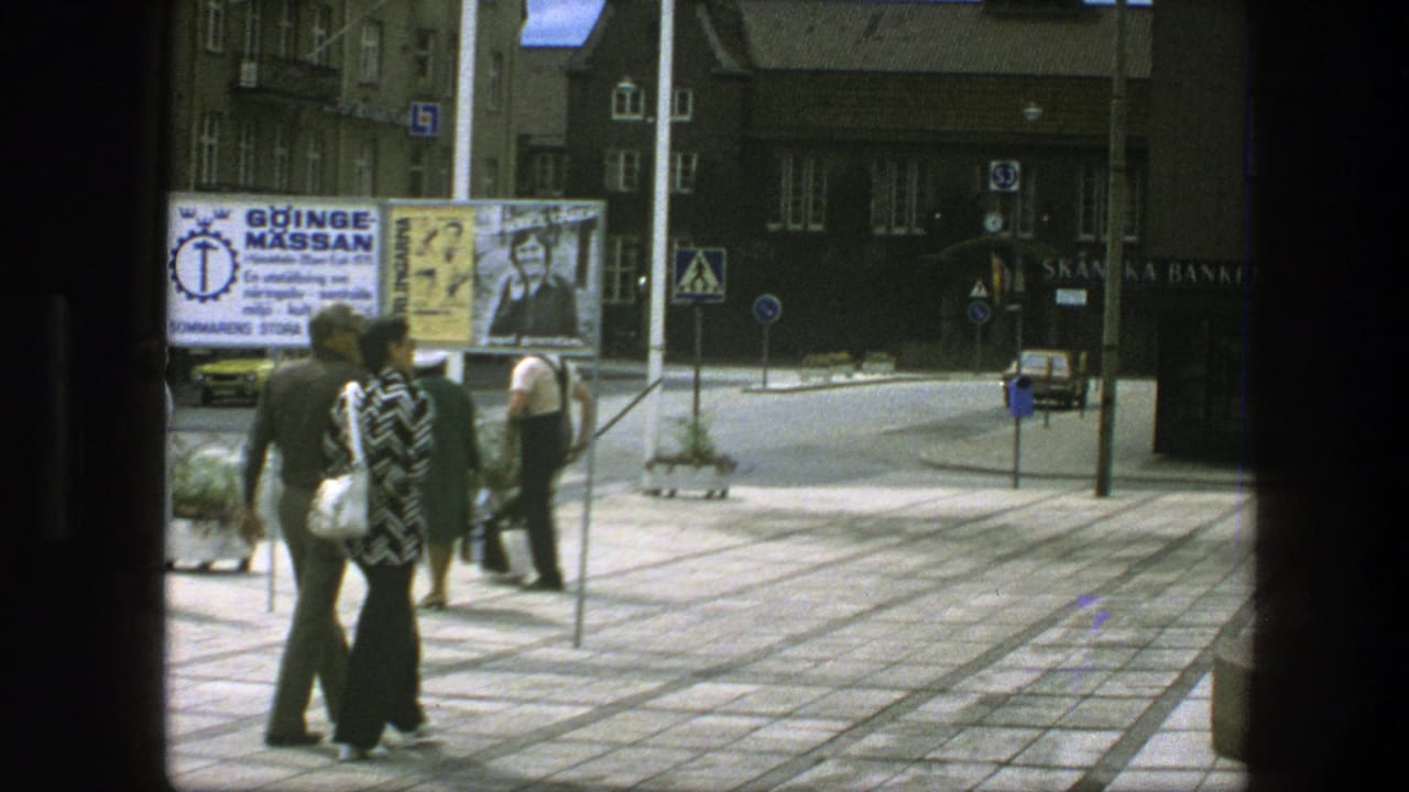 Pedestrians in 1970s Copenhagen walk through a paved plaza, with advertising boards, street signs, and shopfronts visible behind them.