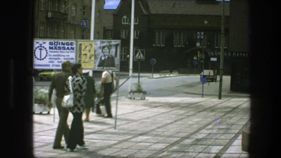 Pedestrians in 1970s Copenhagen walk through a paved plaza, with advertising boards, street signs, and shopfronts visible behind them.