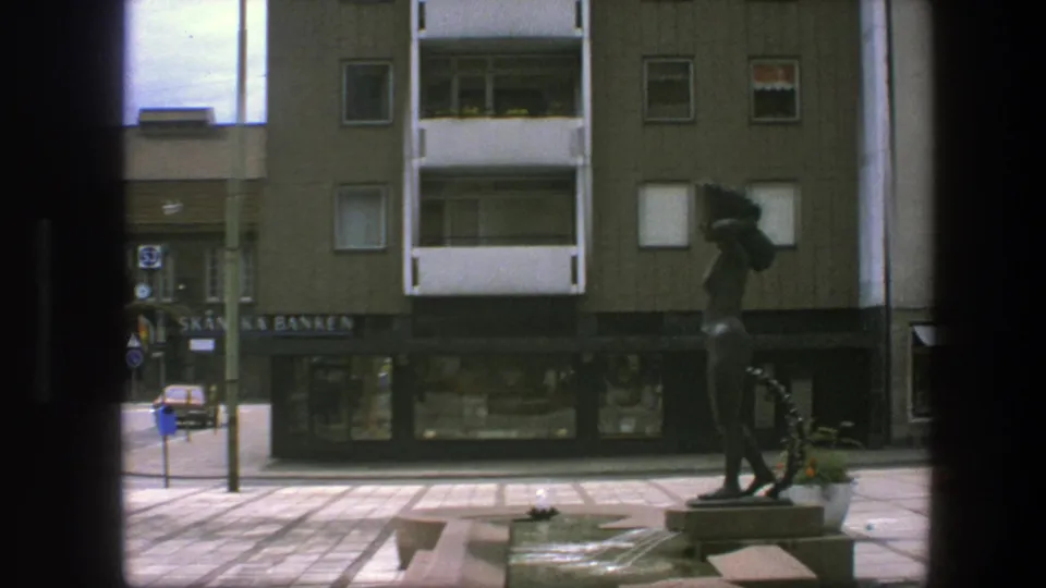 A dark statue stands on a paved Copenhagen plaza in front of apartment balconies, shop windows, and Skandinaviska Banken signage.