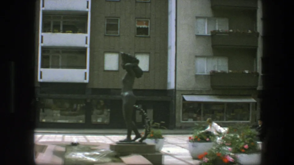The Copenhagen plaza view shows a dark statue, flower planters, shopfronts, and apartment balconies in daylight.