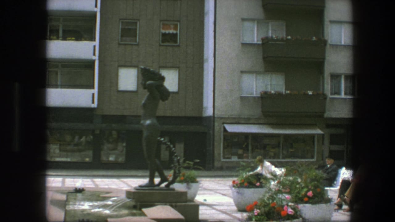 Bronze statue, flower planters, benches, and apartment buildings around a paved square.