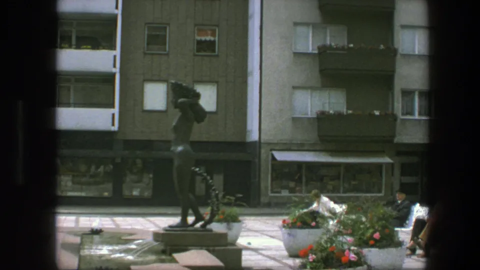 1975 Copenhagen public square with a bronze statue, planted flowers, benches, shop windows, and apartment balconies in soft 8mm color.
