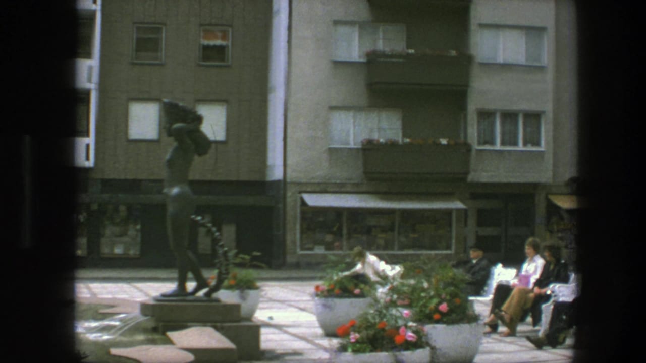 People sit on benches near a statue and flower planters in front of apartment buildings.