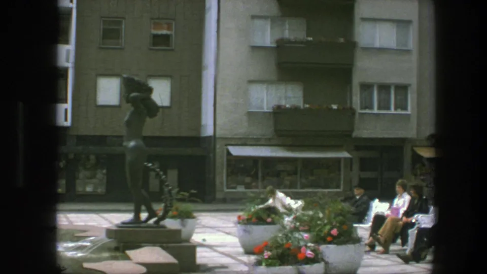 Adults relax on benches near a bronze statue and flower planters in a Copenhagen square, with apartments and shops behind them.