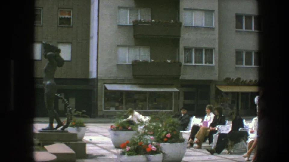 A Copenhagen public square in 1975 shows a statue, bright flower planters, and adults seated on benches below residential balconies.