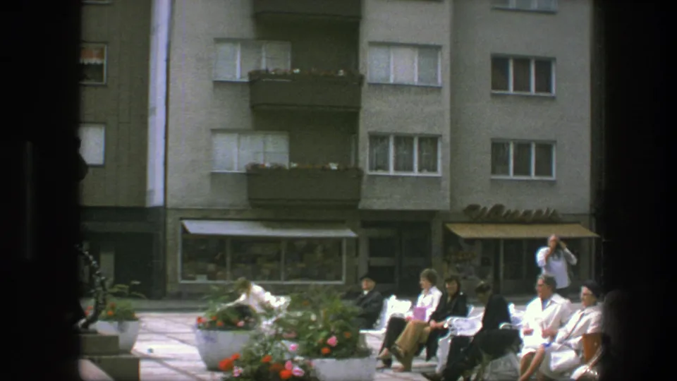 Several adults gather around benches and flower planters in a Copenhagen square, with storefront awnings and apartment windows in the background.