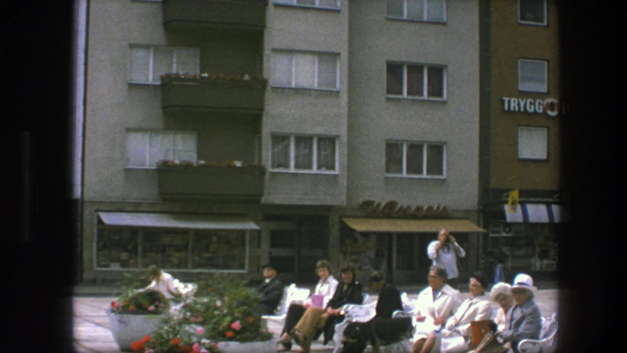 Adults sit on white benches in a sunny plaza beneath apartment buildings and shop awnings.