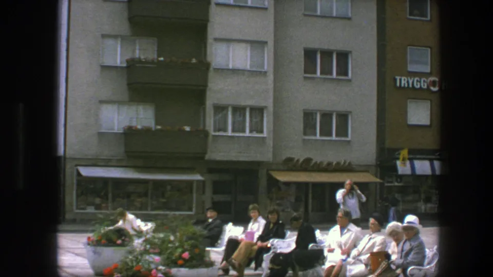 1975 Copenhagen plaza view with adults seated on white benches, flower planters, apartment facades, storefront awnings, and partial Trygg Hansa signage.
