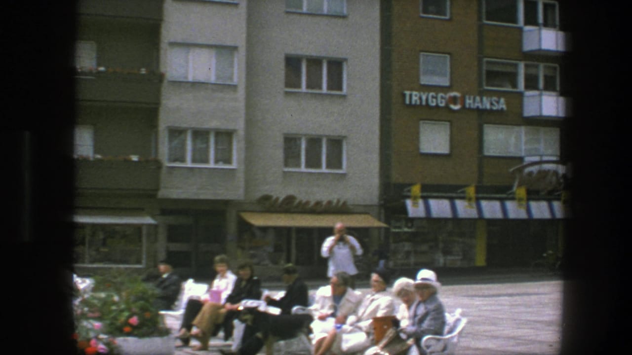 A small group sits on benches while a standing man appears to hold a camera near storefronts.