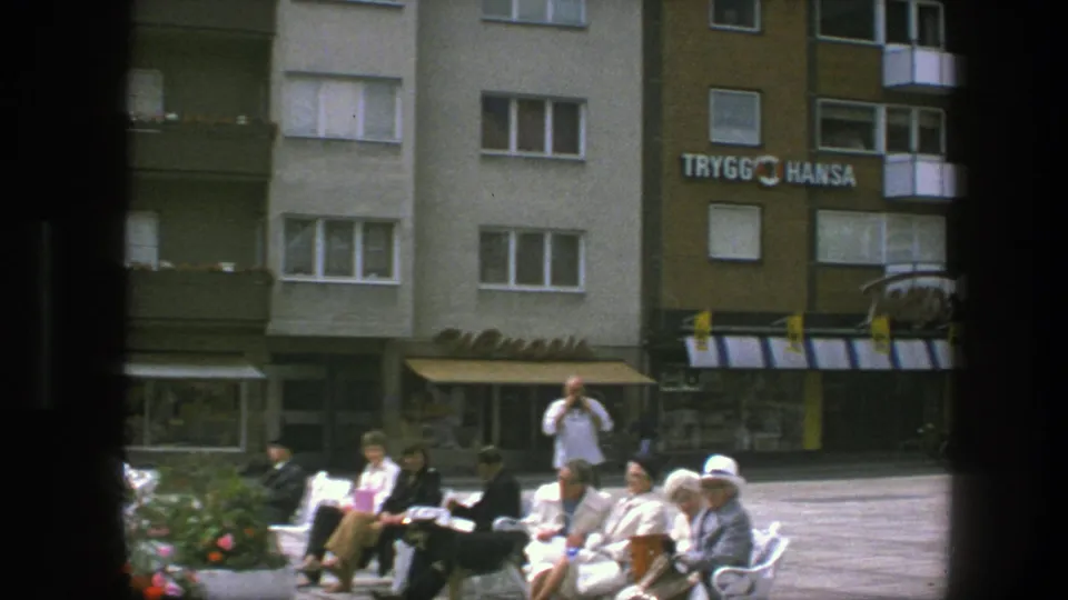 1975 Copenhagen street scene with seated adults on plaza benches, a man possibly taking a photo, striped awnings, balconies, and Trygg Hansa signage.