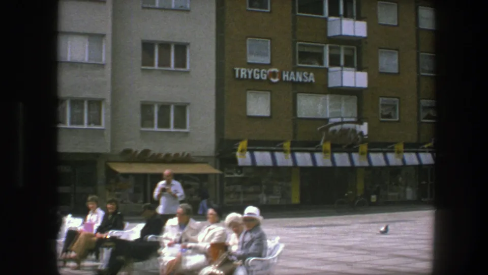 1975 Copenhagen plaza frame showing bench seating, several adults, a standing man, mid-century apartments, awnings, and prominent Trygg Hansa lettering.