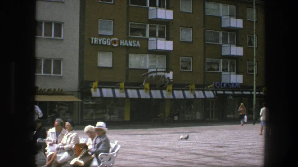 1975 Copenhagen plaza view with adults on benches, a pigeon on the paving, pedestrians at the edge, balconies, awnings, and visible Trygg Hansa signage.
