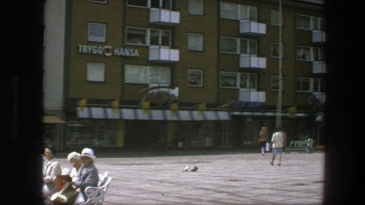 Paved city square with benches, seated adults, doves, and a building marked Trygg Hansa.