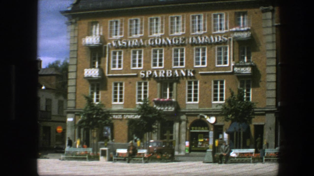 Brick bank building with large Sparbank signage, benches, trees, and pedestrians in a square.
