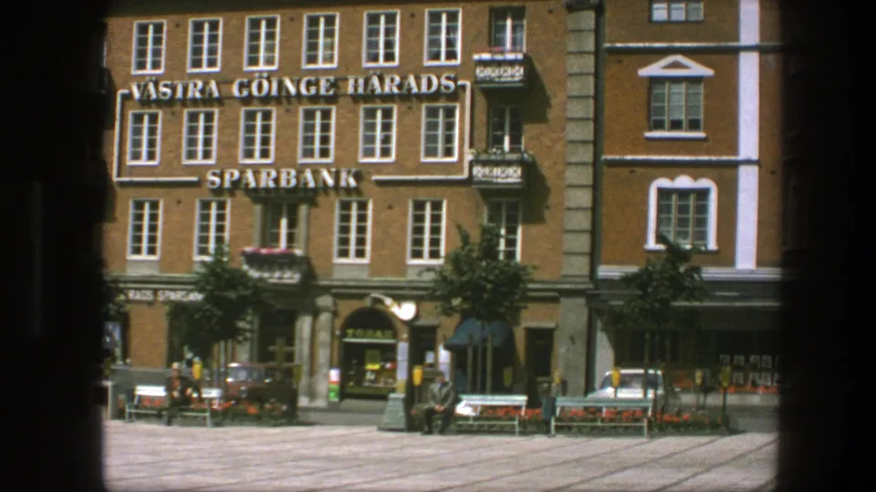 Copenhagen 1975 frame showing the bank building, neighboring storefronts, public benches, small trees, pedestrians, and parked vintage cars.