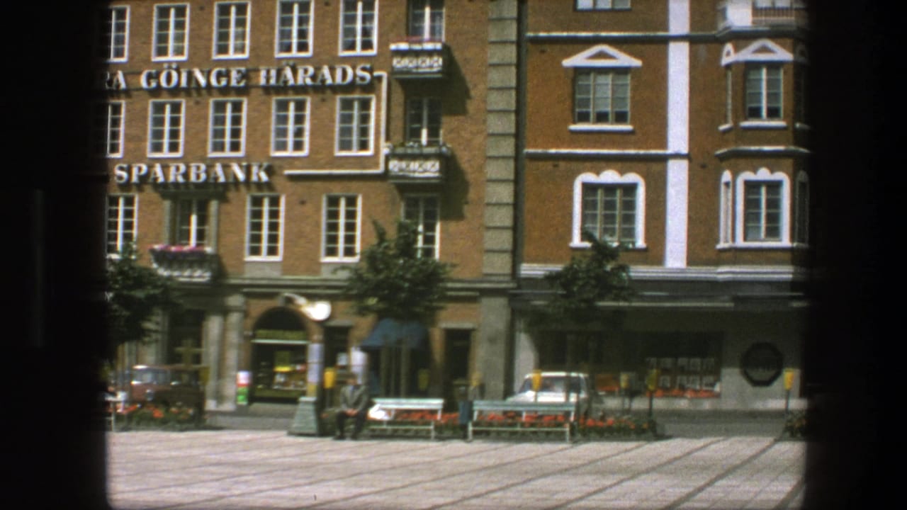 1975 archival 8mm view of a Scandinavian square, showing a brick building with 'SPARBANK' lettering, benches, flowerbeds and people near storefronts.