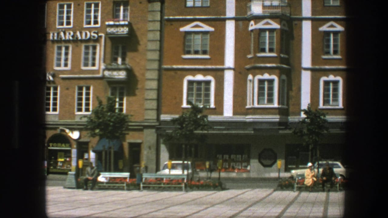 Archival 8mm frame from the 1975 Copenhagen clip showing a quiet square, bank signage, parked car, benches, flowerbeds and pedestrians.