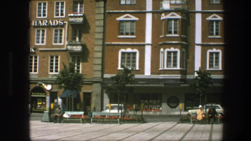 Archival 8mm frame from the 1975 Copenhagen clip showing a quiet square, bank signage, parked car, benches, flowerbeds and pedestrians.