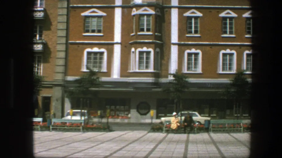 1975 8mm street view with brick upper floors, shop windows, a parked car, benches, flowerbeds and a person crossing the square.