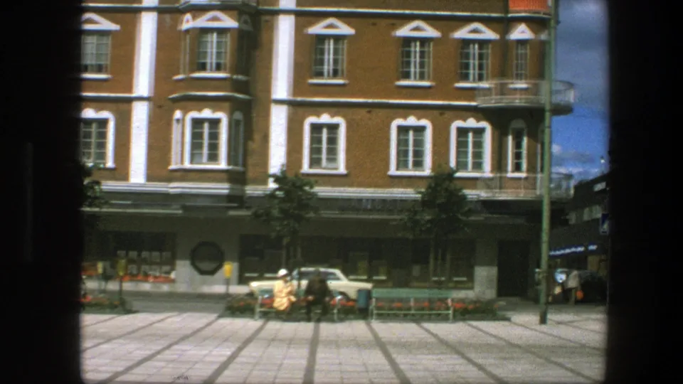 Archival 8mm frame showing the square edge, storefront facade, benches, flowerbeds, pedestrians, a parked car and a vertical street pole.