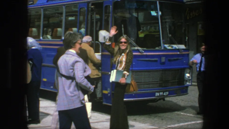 1975 Copenhagen bus-stop scene with a blue public bus, passengers boarding, and a woman in sunglasses waving at the camera.