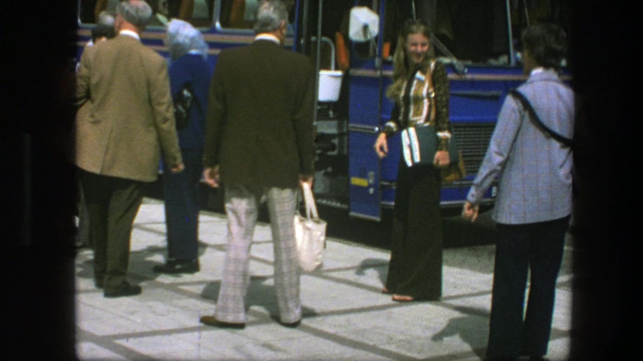 Adults stand near a blue bus on a sidewalk, including a woman in flared pants and a man carrying a white handbag.
